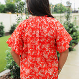 Woman with dark hair standing in a back profile position wearing the Bell Top in a lifestyle scene. The focal point of this image is the white floral print against a red background, ruffled sleeves and pleating on collar. The model paired this top with light-wash denim jeans. 