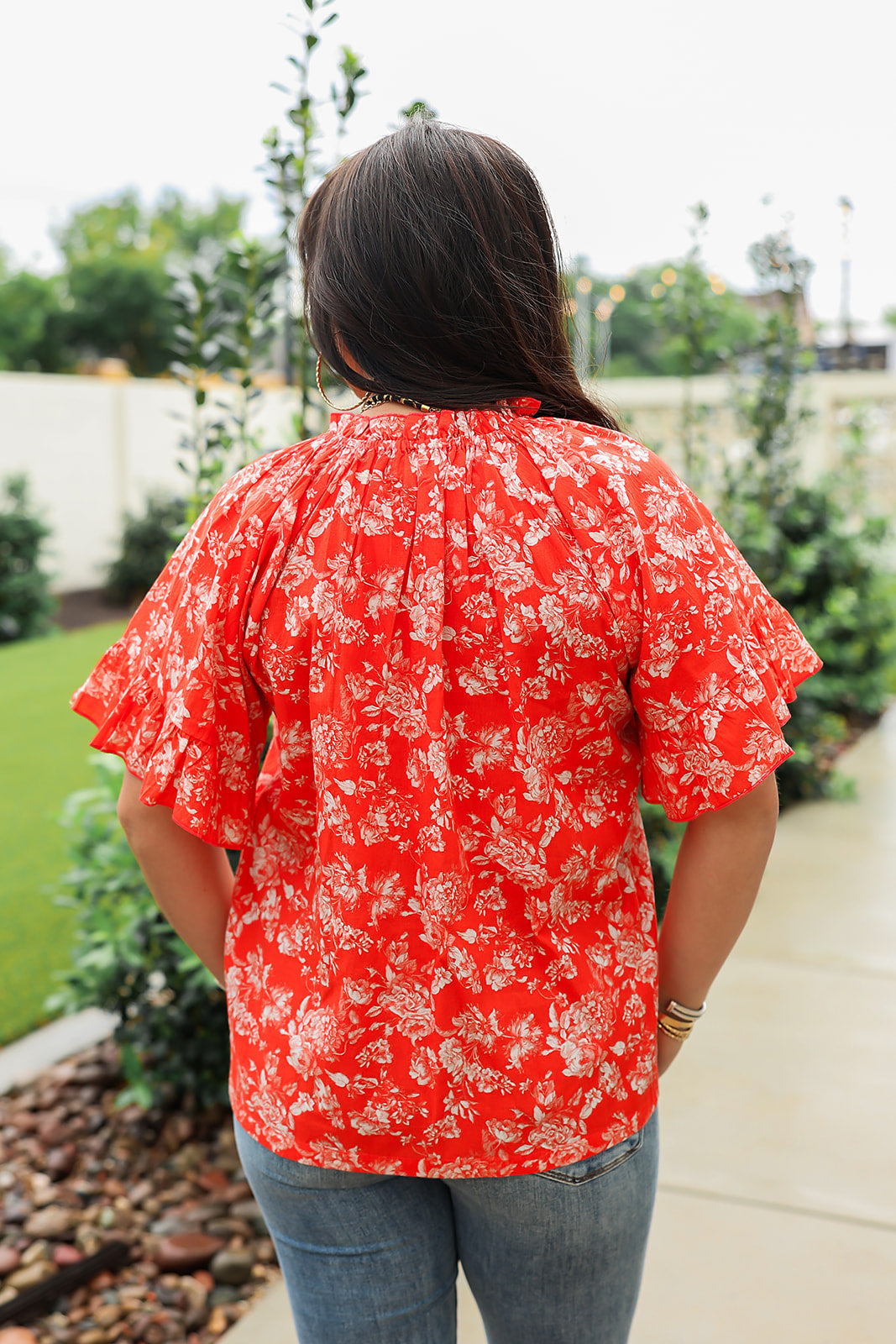 Woman with dark hair standing in a back profile position wearing the Bell Top in a lifestyle scene. The focal point of this image is the white floral print against a red background, ruffled sleeves and pleating on collar. The model paired this top with light-wash denim jeans. 