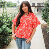 Woman with dark hair standing in a front profile position wearing the Bell Top in a lifestyle scene. The focal point of this image is the floral, tassels, and v-neckline. The model paired this top with light-wash denim jeans. 