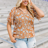Woman with blonde hair standing in a front profile position wearing the Bell Top at a football field. The focal point of this image is the front graphics, tassels, mid-ranged collar and v-neckline. The model paired this top with light-wash denim shorts.