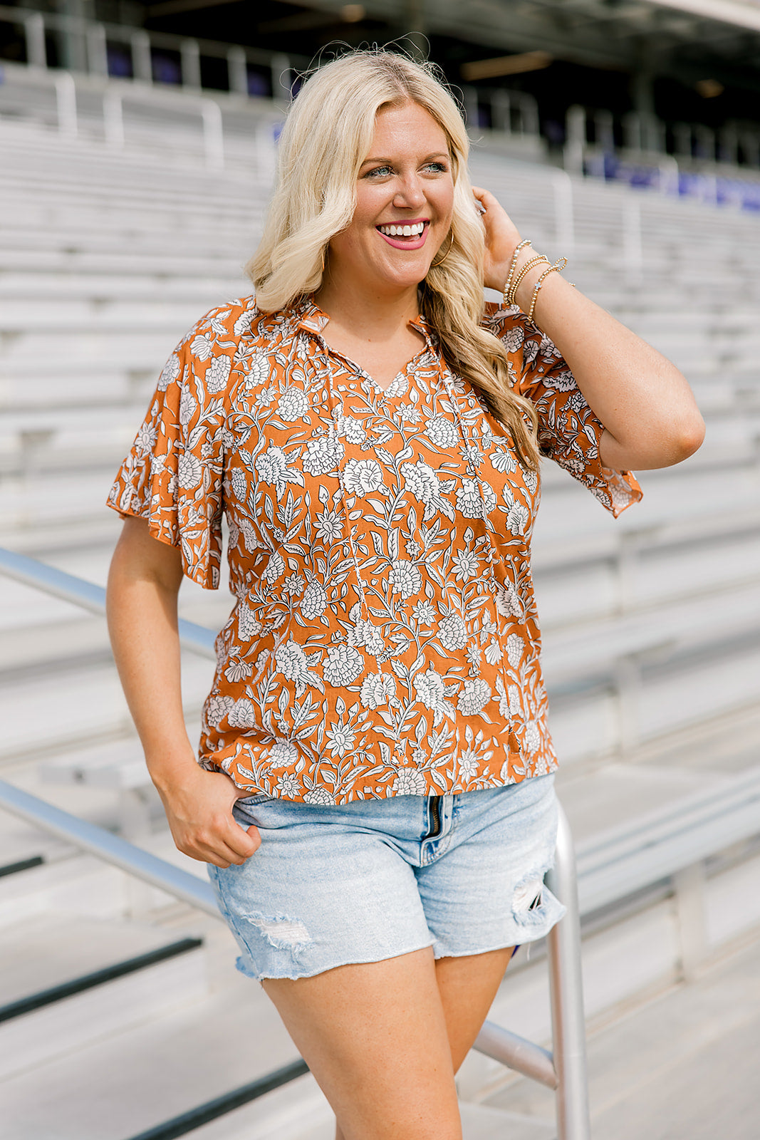 Woman with blonde hair standing in a front profile position wearing the Bell Top at a football field. The focal point of this image is the front graphics, tassels, mid-ranged collar and v-neckline. The model paired this top with light-wash denim shorts.