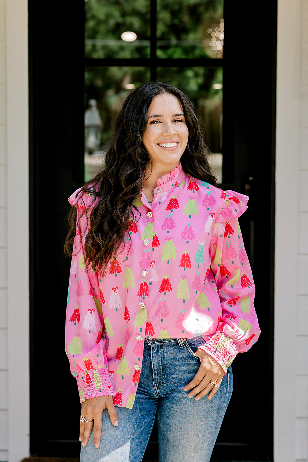 Woman with dark hair wearing a multi-color Christmas tree graphic long-sleeve with ruffles on sleeves. Model paired this top with dark-wash denim jeans. 