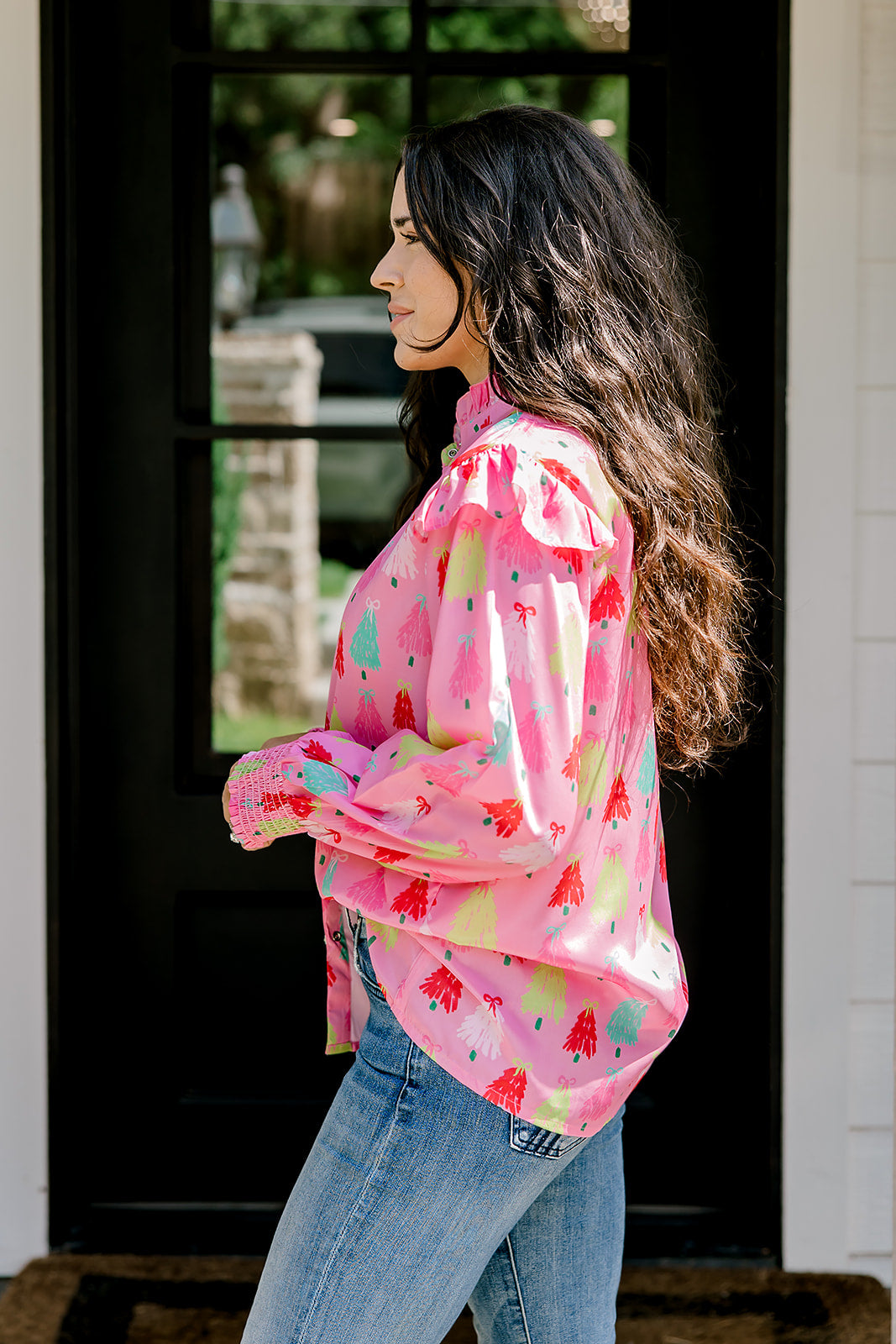 Woman with dark hair wearing a multi-color Christmas tree graphic long-sleeve with ruffles on sleeves. Model paired this top with dark-wash denim jeans. 