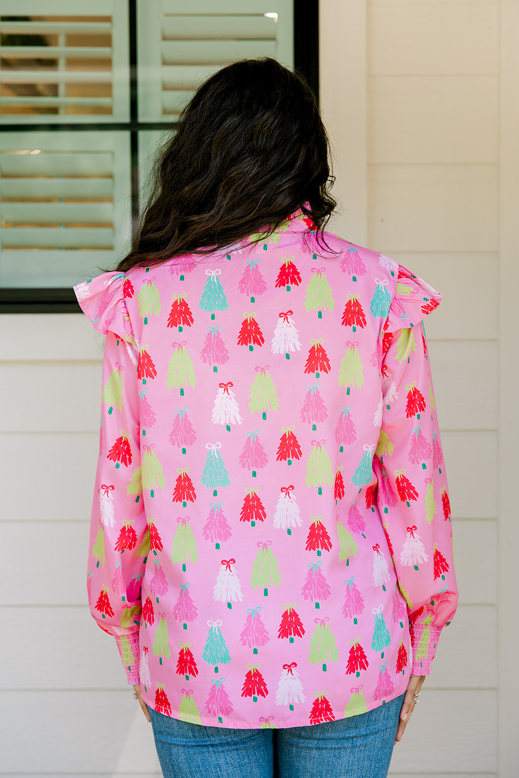 Woman with dark hair wearing a multi-color Christmas tree graphic long-sleeve with ruffles on sleeves. Model paired this top with dark-wash denim jeans. 