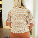 Woman with blonde hair standing in a back profile position wearing the Brinley Top in a lifestyle scene. The focal point of this image is the gingham pattern and patch-work embroidery on ruffled sleeves. The model paired this top with a burnt orange skirt. 
