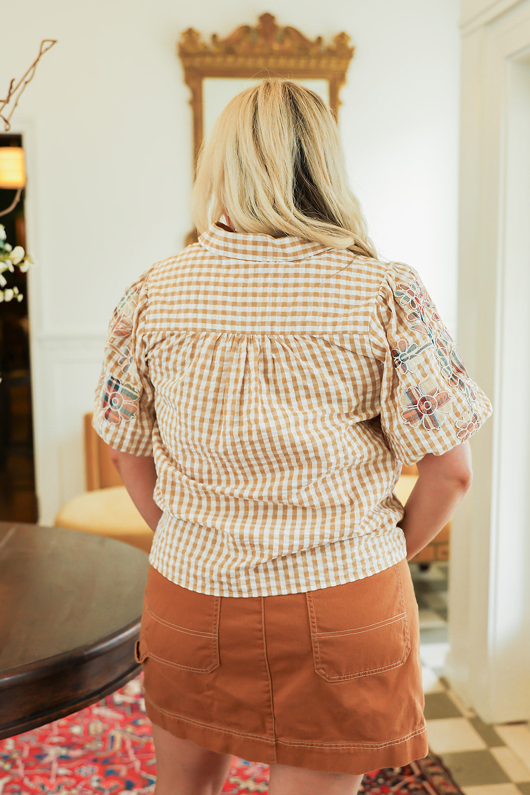 Woman with blonde hair standing in a back profile position wearing the Brinley Top in a lifestyle scene. The focal point of this image is the gingham pattern and patch-work embroidery on ruffled sleeves. The model paired this top with a burnt orange skirt. 