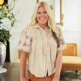 Woman with blonde hair standing in a front profile position wearing the Brinley Top in a lifestyle scene. The focal point of this image is the front embroidered detailing on top as well as the buttons down the front and embroidered sleeves. The model paired this top with a burnt orange skirt. 