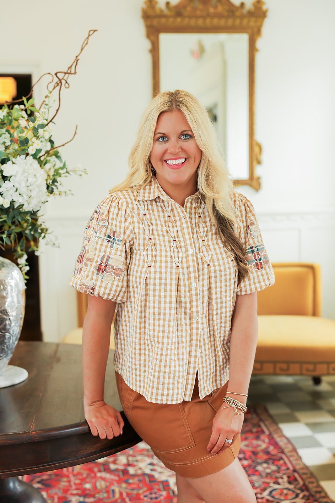 Woman with blonde hair standing in a front profile position wearing the Brinley Top in a lifestyle scene. The focal point of this image is the front embroidered detailing on top as well as the buttons down the front and embroidered sleeves. The model paired this top with a burnt orange skirt. 