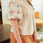 Woman with blonde hair standing in a side profile position wearing the Brinley Top in a lifestyle scene. The focal point of this image is the patch-work embroidered ruffled sleeves. The model paired this top with a burnt orange skirt. 