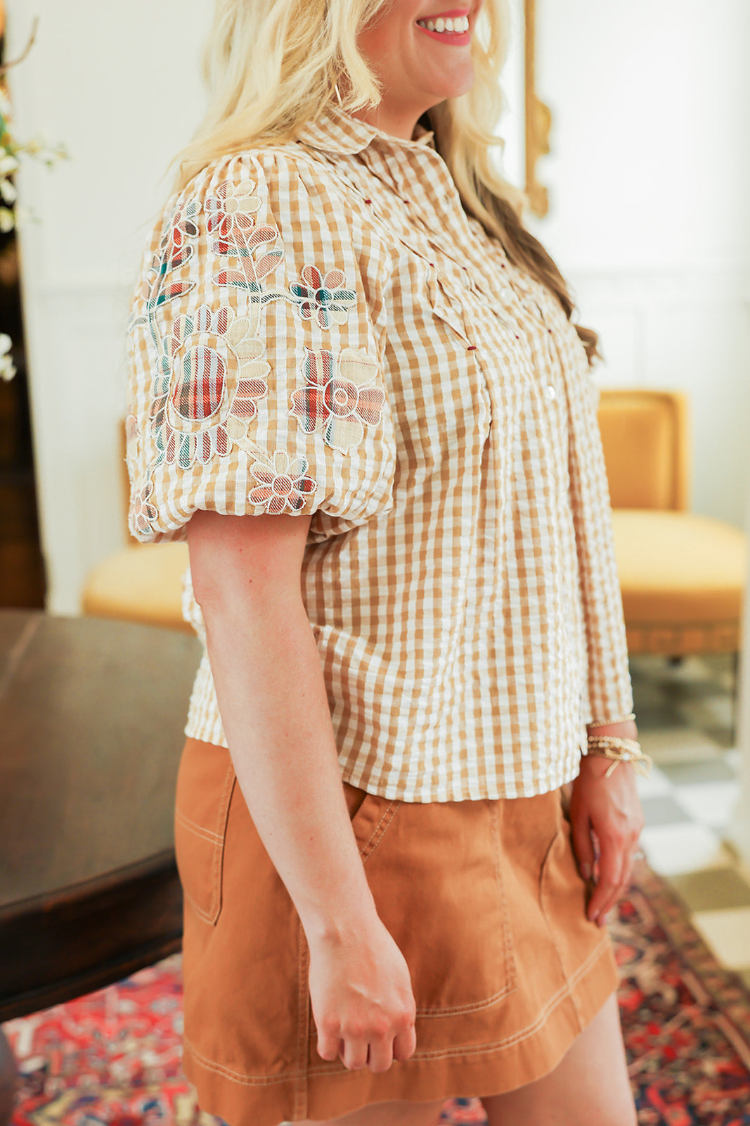 Woman with blonde hair standing in a side profile position wearing the Brinley Top in a lifestyle scene. The focal point of this image is the patch-work embroidered ruffled sleeves. The model paired this top with a burnt orange skirt. 