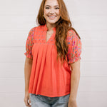 Woman wearing a coral blouse with floral sleeves against a white background facing front. 