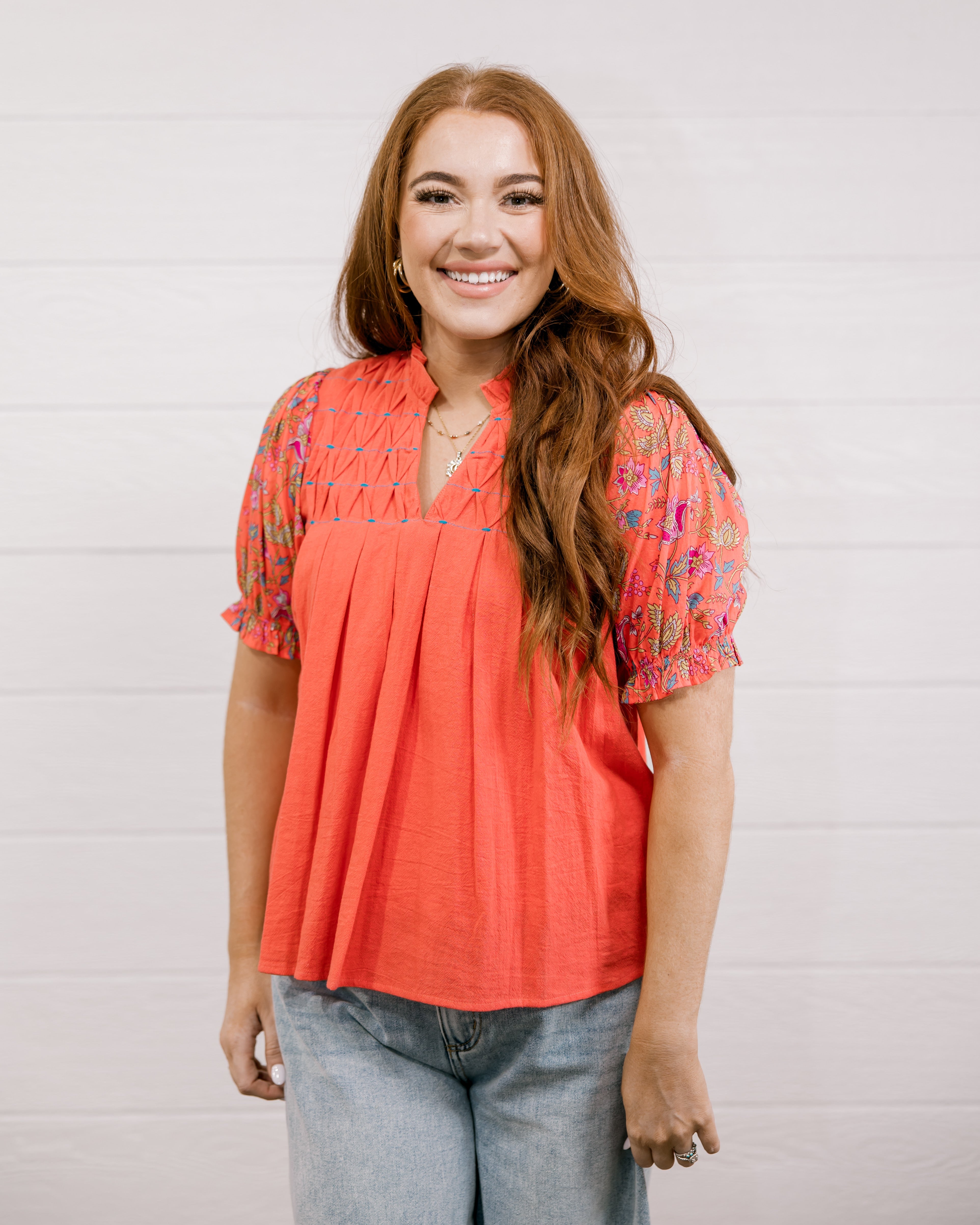 Woman wearing a coral blouse with floral sleeves against a white background facing front. 