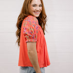 Woman wearing a coral floral blouse against a light background facing side.