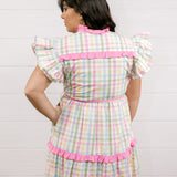 Woman wearing a checkered dress with pink accents against a white background facing back. 