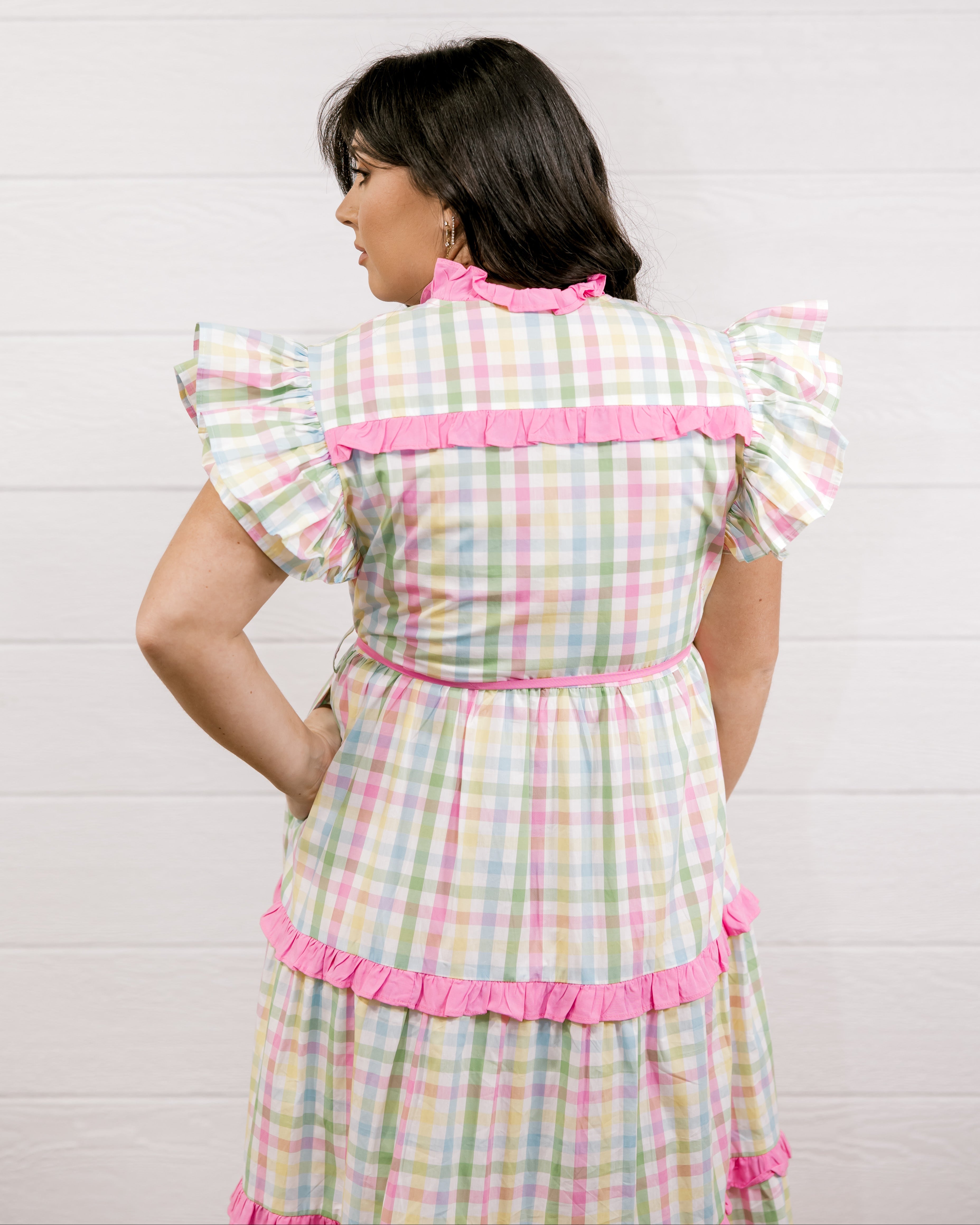 Woman wearing a checkered dress with pink accents against a white background facing back. 