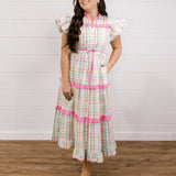 Woman wearing a checkered dress with pink accents against a white wall facing the front. 