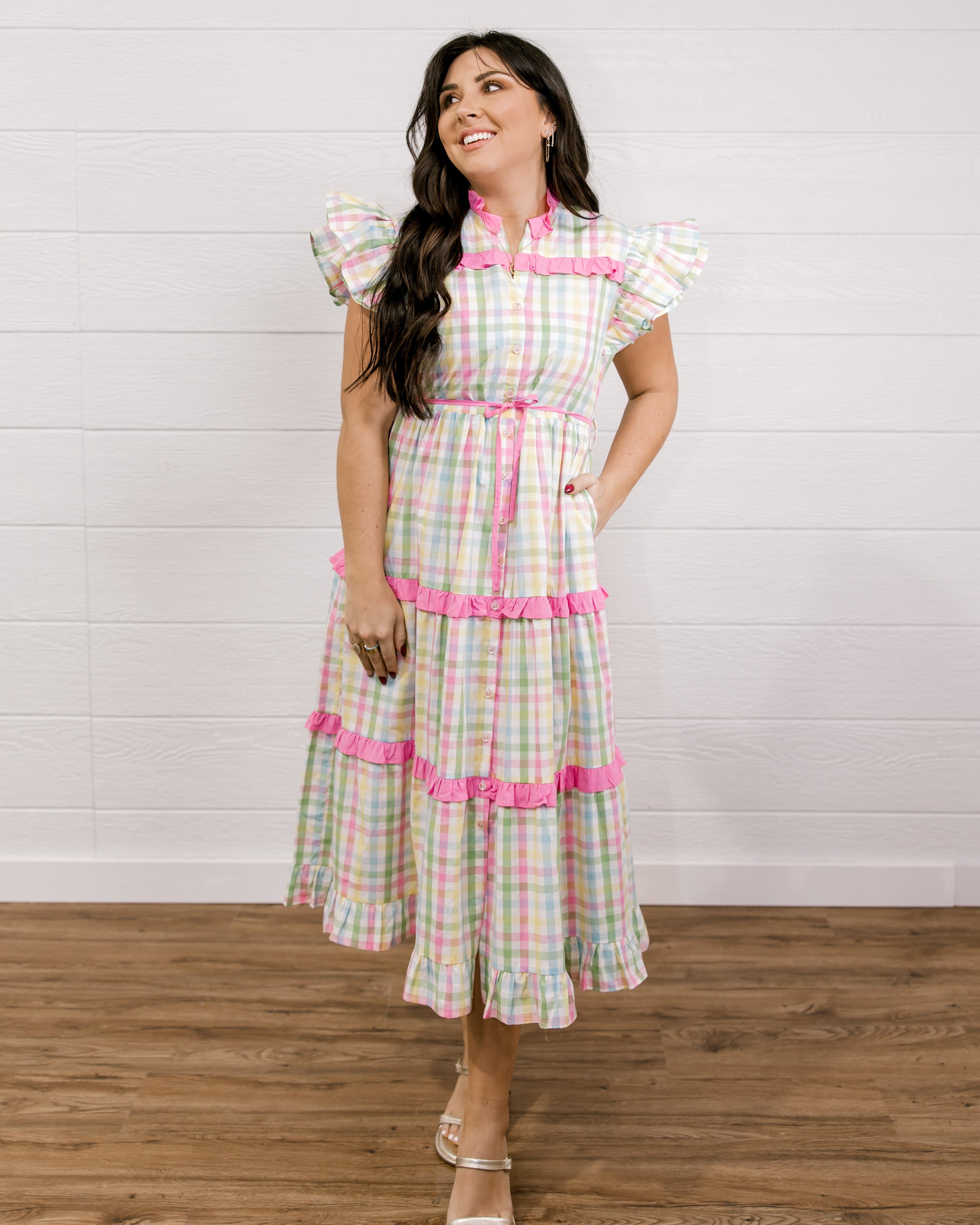 Woman wearing a checkered dress with pink accents against a white wall facing the front. 