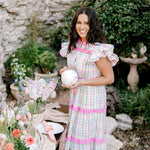 Woman in a pastel dress holding a white ball in an outdoor setting with flowers and a stone wall.