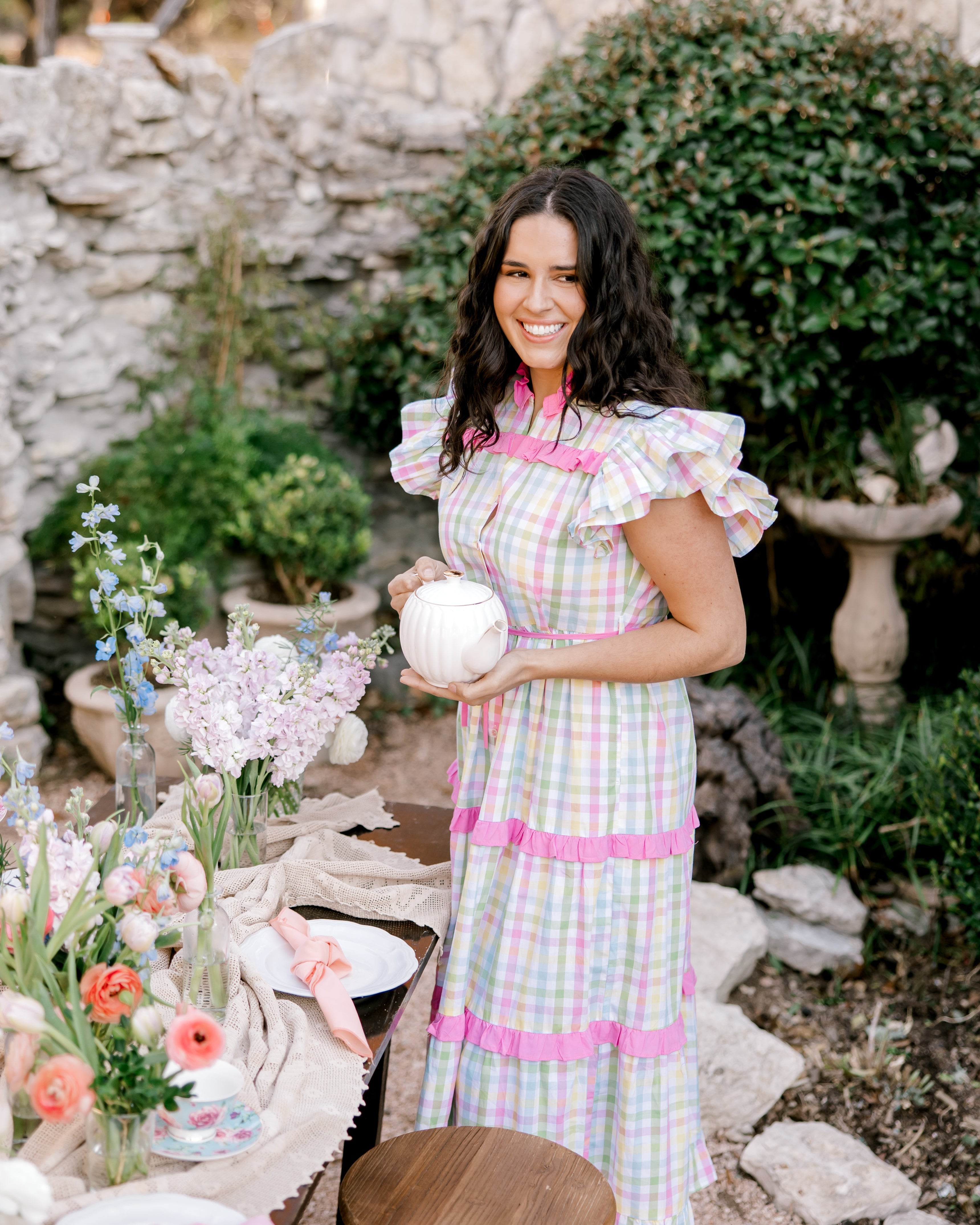 Woman in a pastel dress holding a white ball in an outdoor setting with flowers and a stone wall.
