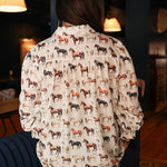 Woman with dark hair standing in a back profile position wearing the Caroline Top in a lifestyle scene. The focal point of this image is the multi-color horse graphic on the back. The model paired this top with dark-wash denim jeans. 