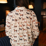 Woman with dark hair standing in a back profile position wearing the Caroline Top in a lifestyle scene. The focal point of this image is the multi-color horse graphic on the back. The model paired this top with dark-wash denim jeans. 