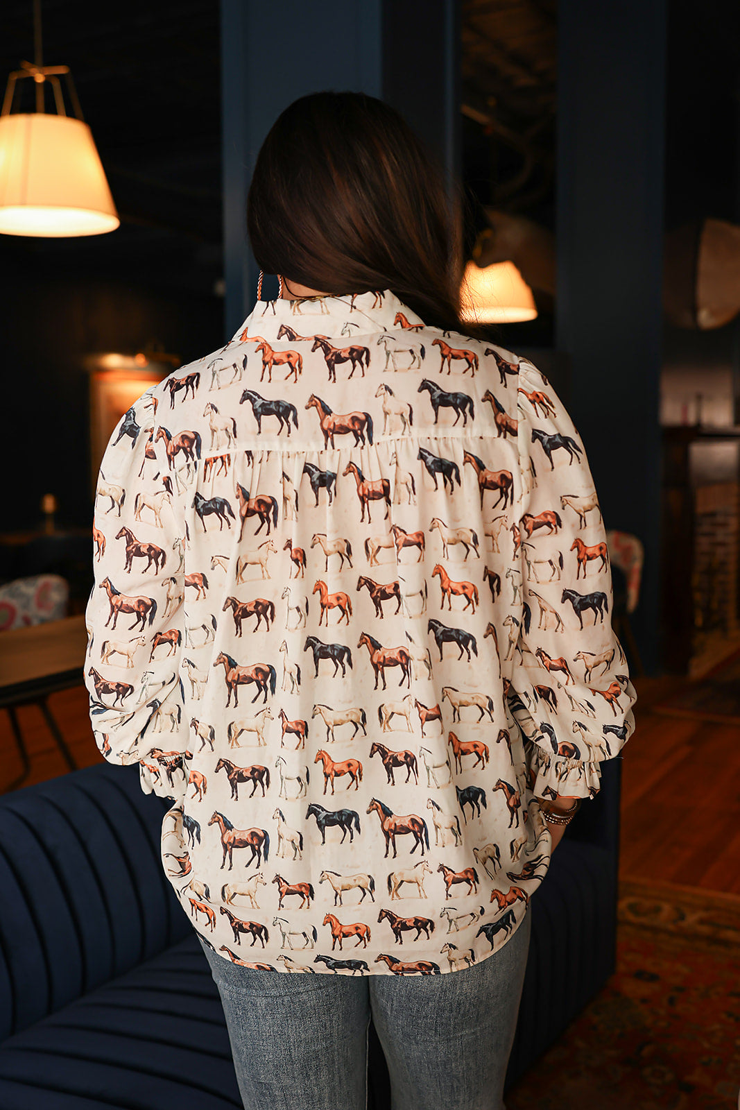 Woman with dark hair standing in a back profile position wearing the Caroline Top in a lifestyle scene. The focal point of this image is the multi-color horse graphic on the back. The model paired this top with dark-wash denim jeans. 