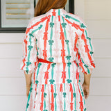 Woman with red hair wearing a long-sleeve dress with pink, red, and green graphic bows. Dress comes with red-fabric that is tied in front. 