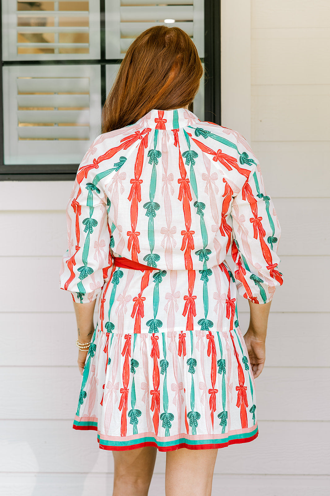 Woman with red hair wearing a long-sleeve dress with pink, red, and green graphic bows. Dress comes with red-fabric that is tied in front. 