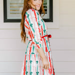 Woman with red hair wearing a long-sleeve dress with pink, red, and green graphic bows. Dress comes with red-fabric that is tied in front. 