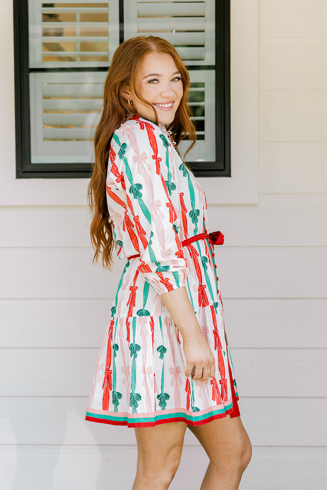 Woman with red hair wearing a long-sleeve dress with pink, red, and green graphic bows. Dress comes with red-fabric that is tied in front. 