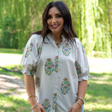 Woman with dark hair wearing the Courtney Top in a front facing position with lifestyle background. The background  is heavy in greenery, and the model has the top paired with light-wash denim jeans. 
