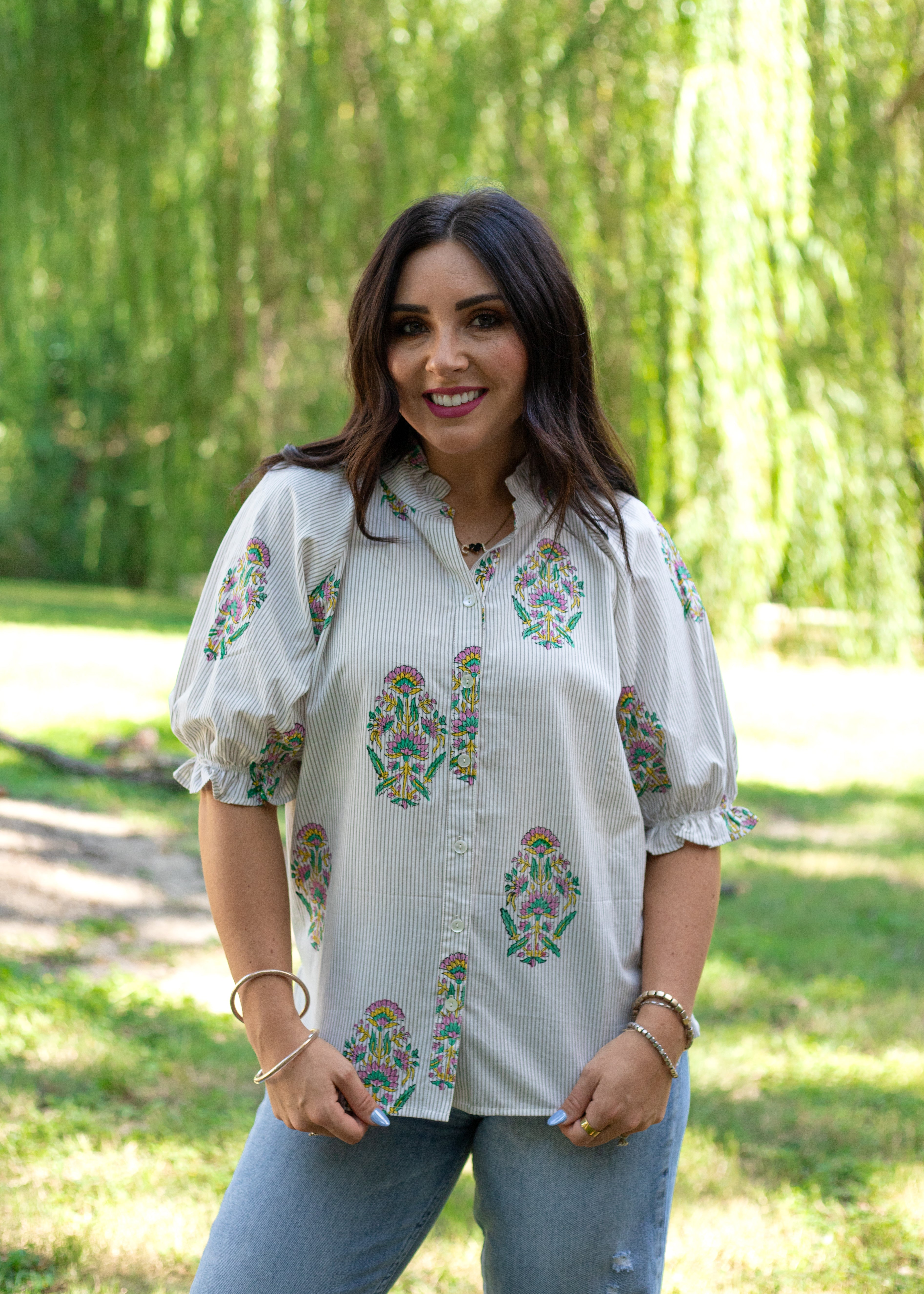 Woman with dark hair wearing the Courtney Top in a front facing position with lifestyle background. The background  is heavy in greenery, and the model has the top paired with light-wash denim jeans. 