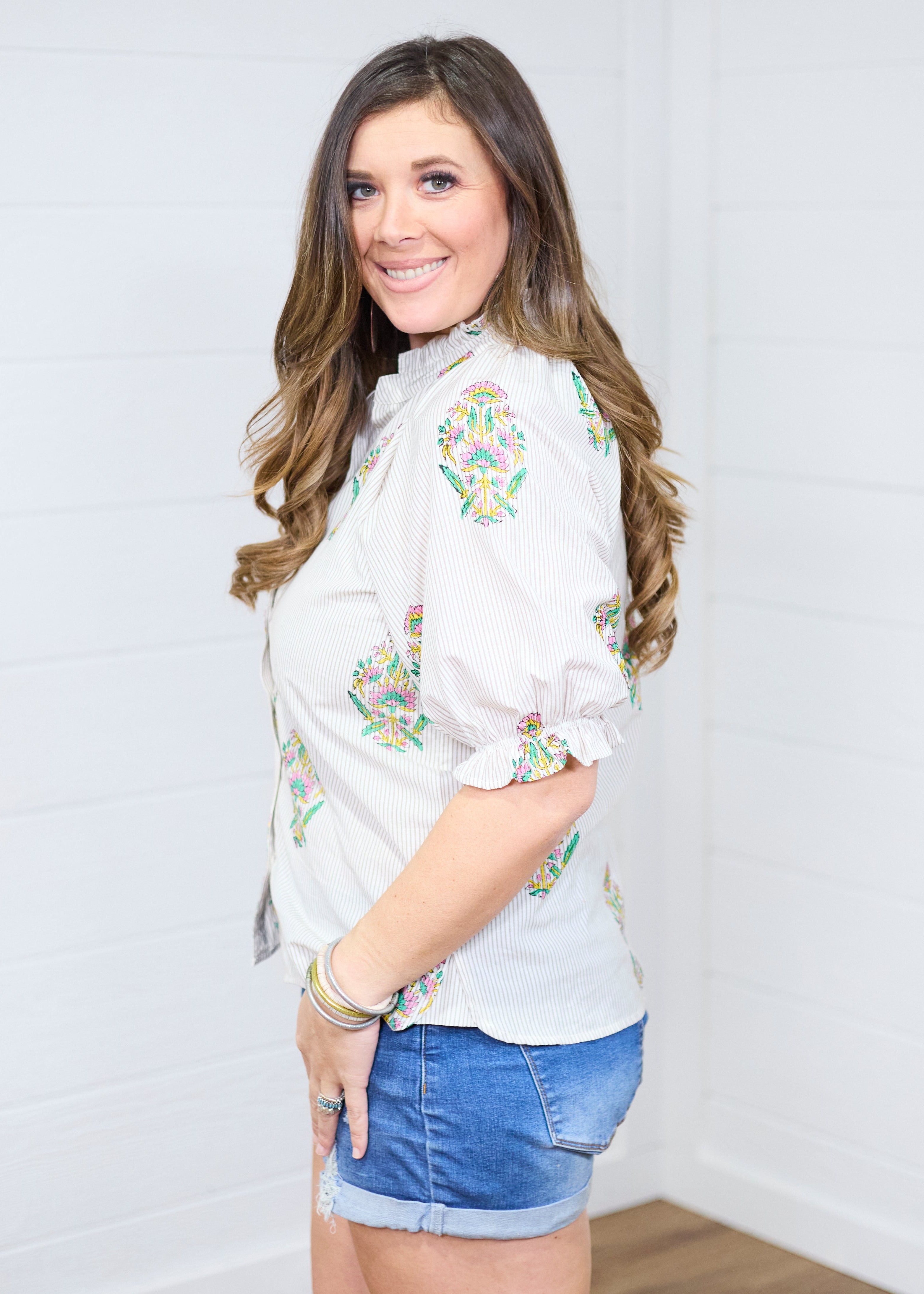 Woman with dark hair in a side profile position with the Courtney Top against a white background. This image is showing the side profile and sleeves of top. She has the top paired with dark-wash denim shorts. 