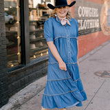 Woman in a blue dress and cowboy hat standing on a sidewalk with a store in the background.