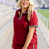 Woman with blonde hair standing in a side profile position wearing the Daisy Top & Jade Skirt in a lifestyle scene. The focal point of this image is the striping along quarter zip, cuff and side of skirt as well as the grooved maroon fabric. 