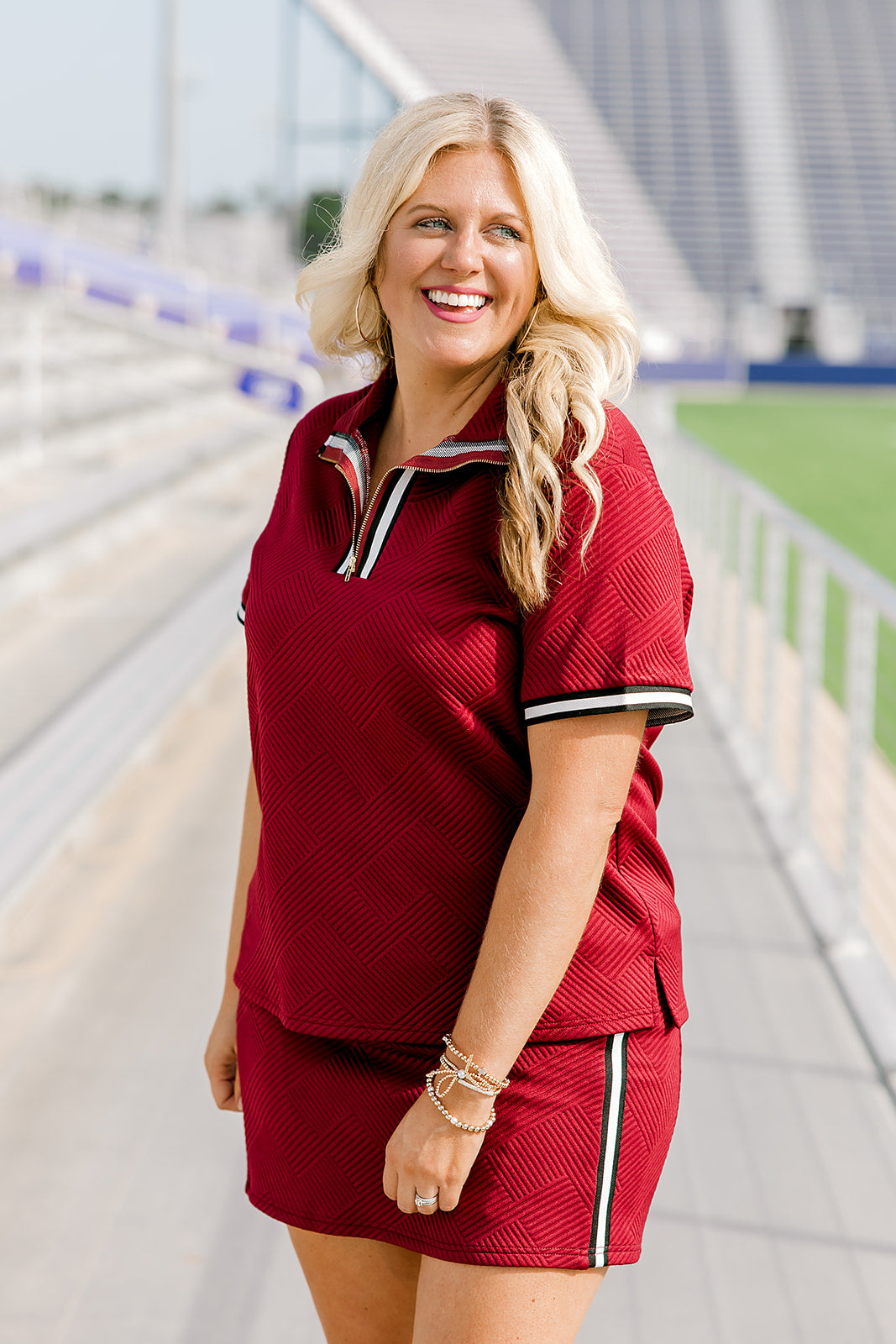 Woman with blonde hair standing in a side profile position wearing the Daisy Top & Jade Skirt in a lifestyle scene. The focal point of this image is the striping along quarter zip, cuff and side of skirt as well as the grooved maroon fabric. 