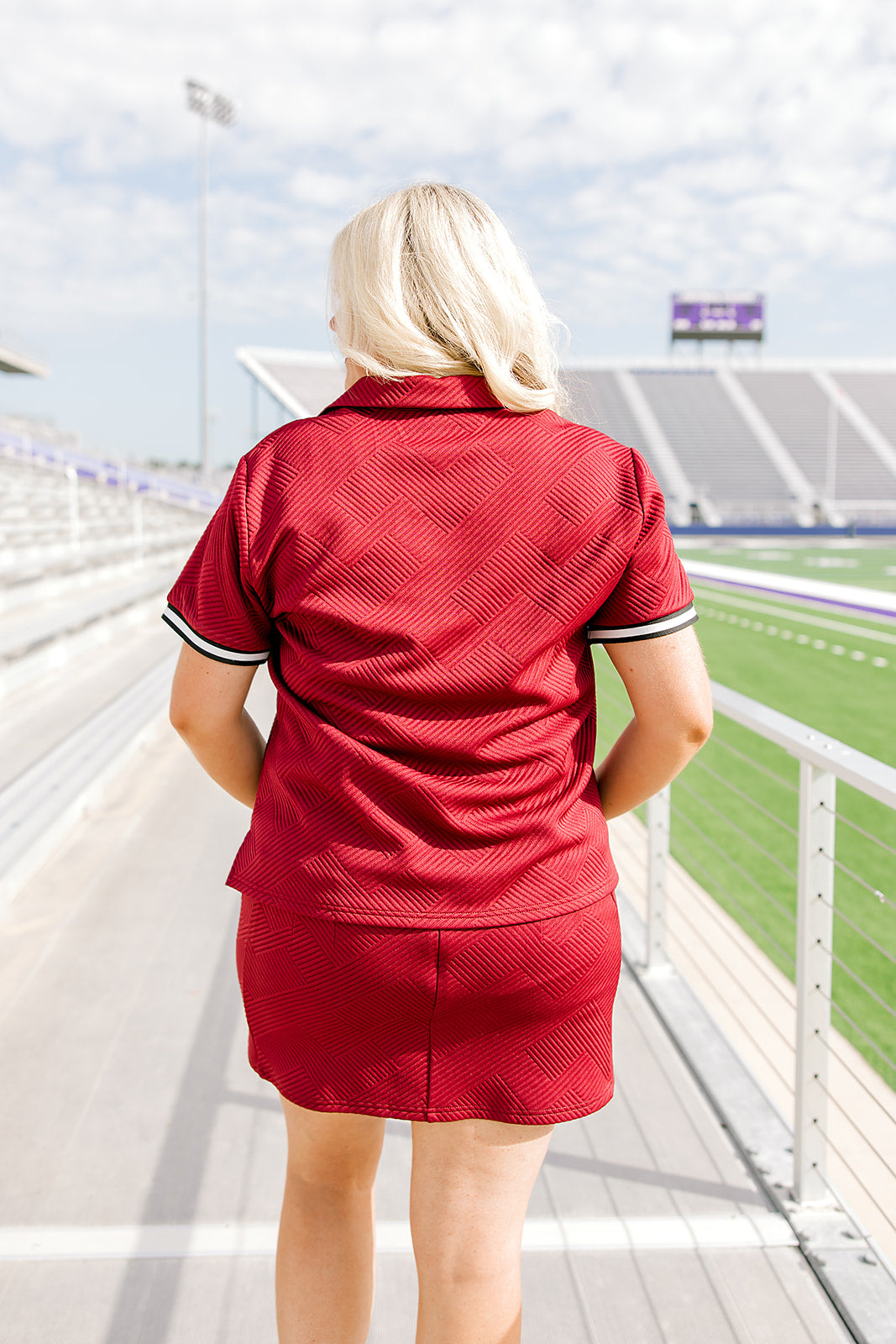 Woman with blonde hair standing in a back profile position wearing the Daisy Top and Jade Skirt at a football field. The focal point of this image is the intricate fabric detail on the back of top and skirt as well as the striping on cuff. 