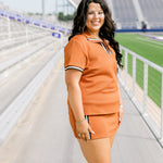 Woman with dark hair standing in a side profile position wearing the Daisy Top and Jane Skirt at a football field. The focal point of this image is to see the length of skirt as well as striping along cuff and down skirt. 
