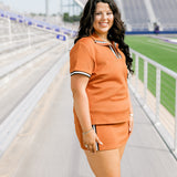 Woman with dark hair standing in a side profile position wearing the Daisy Top and Jane Skirt at a football field. The focal point of this image is to see the length of skirt as well as striping along cuff and down skirt. 