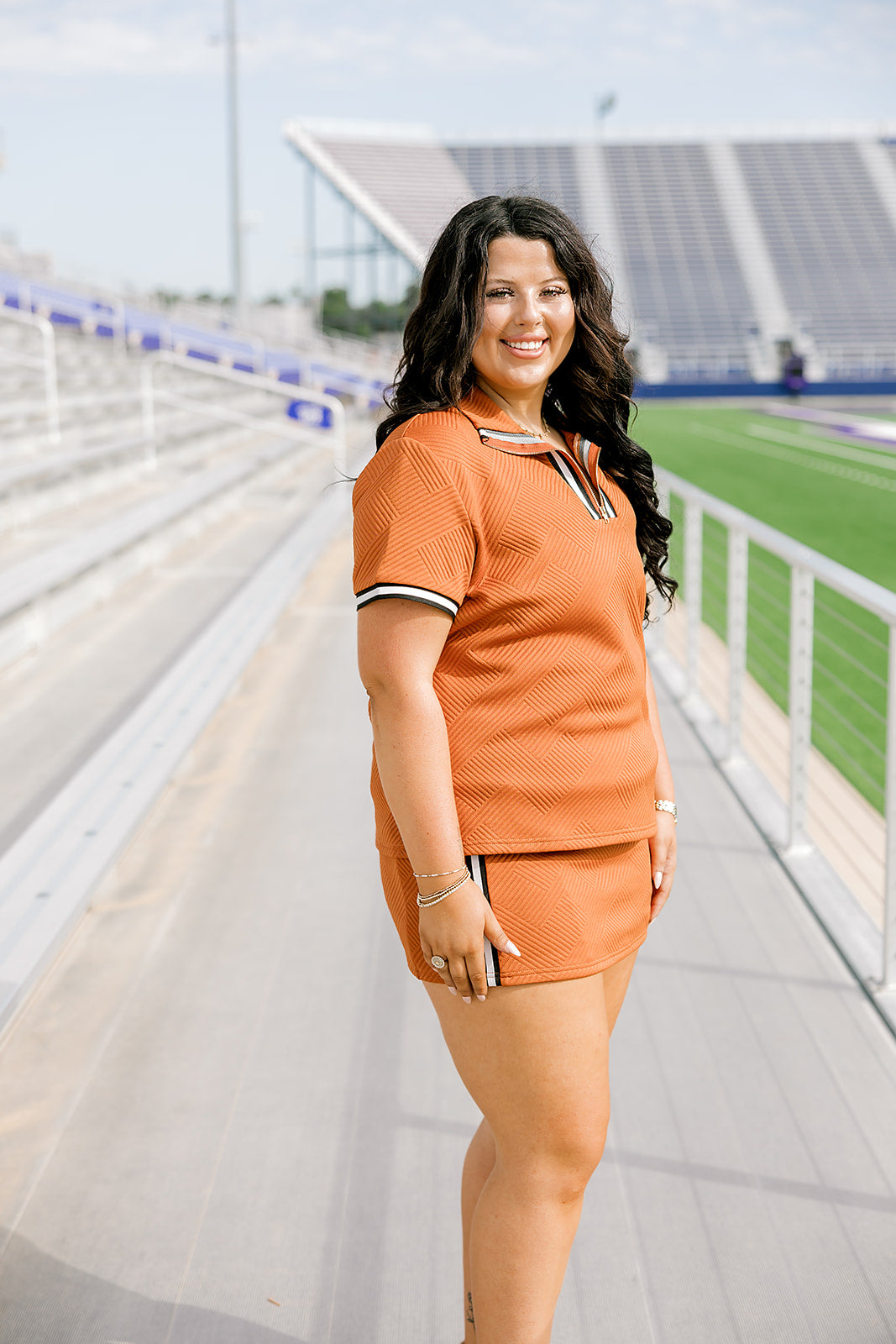 Woman with dark hair standing in a side profile position wearing the Daisy Top and Jane Skirt at a football field. The focal point of this image is to see the length of skirt as well as striping along cuff and down skirt. 