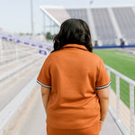 Woman with dark hair standing in a back profile position wearing the Daisy Top and Jane Skirt at a football field. The focal point of this image is the intricate fabric details on back as well as length of skirt and striping along cuff of shirt. 