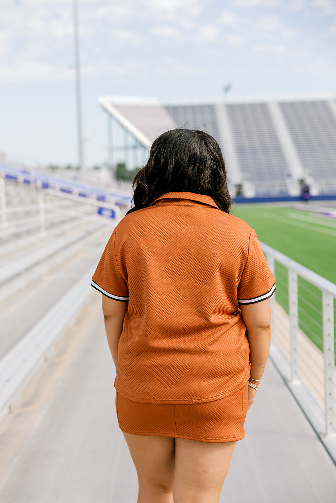 Woman with dark hair standing in a back profile position wearing the Daisy Top and Jane Skirt at a football field. The focal point of this image is the intricate fabric details on back as well as length of skirt and striping along cuff of shirt. 