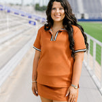 Woman with dark hair standing in a front profile position wearing the Daisy Top and Jane Skirt at a football field. The focal point of this image is the front details of skirt and top as well as striping on the shirt near quarter zip. 