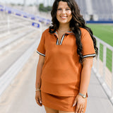 Woman with dark hair standing in a front profile position wearing the Daisy Top and Jane Skirt at a football field. The focal point of this image is the front details of skirt and top as well as striping on the shirt near quarter zip. 