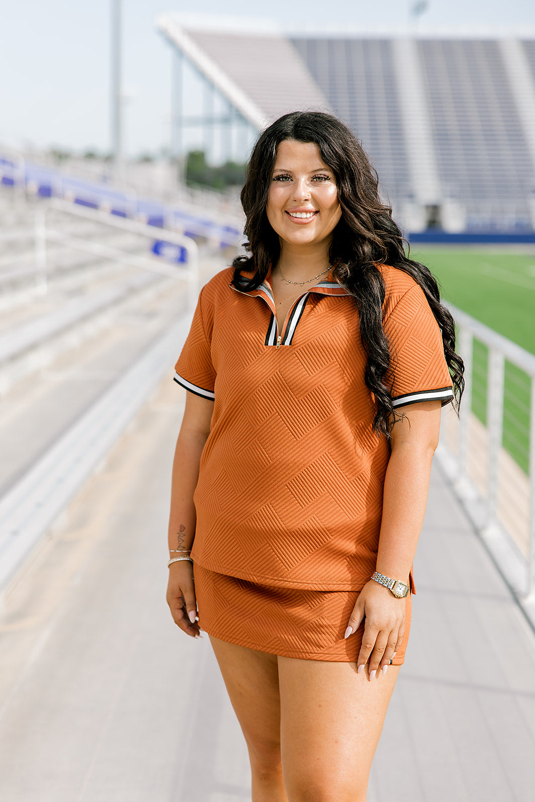 Woman with dark hair standing in a front profile position wearing the Daisy Top and Jane Skirt at a football field. The focal point of this image is the front details of skirt and top as well as striping on the shirt near quarter zip. 