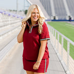 Woman with blonde hair standing in a front profile position wearing the Daisy Top and Jane Skirt at a football field. The focal point of this image is the front details of top and skirt as well as all the areas with striping. Emphasis on the quarter zip with high-collar. 