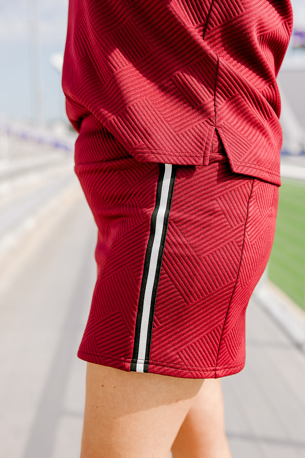 Woman standing in a side profile position wearing the Daisy Top and Jane skirt at a football field. The image is in deep focal length, giving a better look at the pattern detail, striping down side of the skirt as well as length of skirt. 