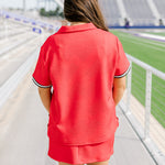 Woman with dark hair standing in a back profile position wearing the Daisy Top and Jane Skirt at a football field. The focal point of this image is the detail of the fabric, length of skirt, and striping along cuff. 