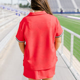 Woman with dark hair standing in a back profile position wearing the Daisy Top and Jane Skirt at a football field. The focal point of this image is the detail of the fabric, length of skirt, and striping along cuff. 