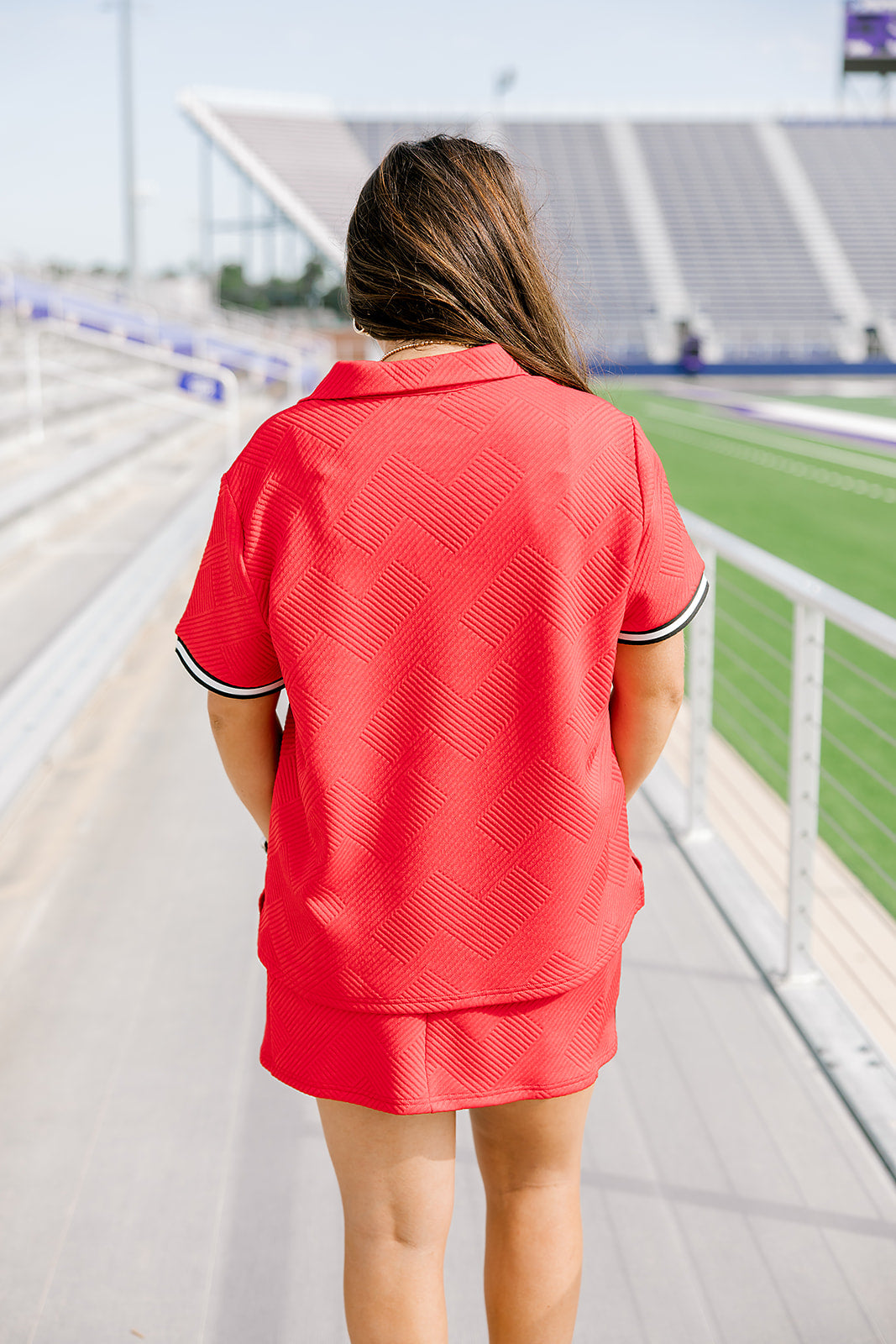 Woman with dark hair standing in a back profile position wearing the Daisy Top and Jane Skirt at a football field. The focal point of this image is the detail of the fabric, length of skirt, and striping along cuff. 